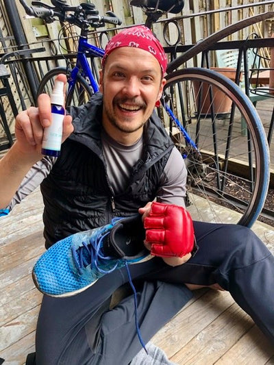 Man with a goatee and a red bandana on his head, sitting in front of a blue bicycle, wearing a kick-boxing glove, holding a running shoe and excitedly showing off his bottle of GOE Spray